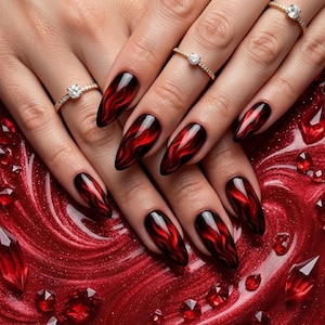 May include: Long, pointed fingernails with a glossy, dark red and black flame design. The nails are decorated with gold rings featuring small, clear gemstones. The background is a textured, red surface with scattered red crystals.