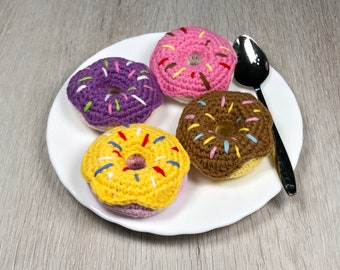 Puede incluir: Cuatro donuts de crochet en un plato blanco. Los donuts son de color rosa, morado, amarillo y marrón con chispas de colores. Una cuchara de plata está en el plato.