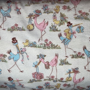 Może przedstawiać: Biały materiał z powtarzającym się wzorem niebieskich i różowych bocianów niosących prezenty. Bociany noszą kapelusze i niosą pluszowe misie, ubranka dla dzieci i pudełka z prezentami. Tło jest jasnoniebieskie z kwiatowym wzorem.