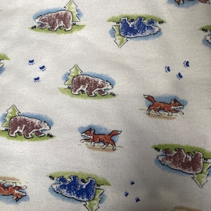 Może przedstawiać: Biały materiał z powtarzającym się wzorem brązowych i niebieskich niedźwiedzi, lisów i sosen. Niedźwiedzie idą, a lisy biegną. Na materiale rozrzucone są również niebieskie ślady łap.