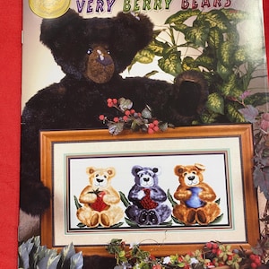May include: Cross stitch pattern booklet featuring three teddy bears holding berries. The booklet cover features a black teddy bear with a green plant in the background. The title of the pattern is "Very Berry Bears".