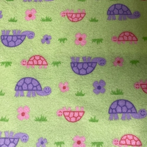 Może przedstawiać: Zielony materiał z powtarzającym się wzorem fioletowych i różowych żółwi i kwiatów. Żółwie są zwrócone w prawo i mają uśmiechnięte twarze. Kwiaty są różowe i fioletowe.