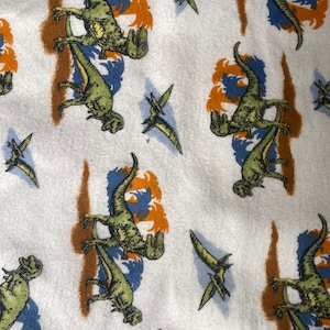 Może przedstawiać: Biały materiał z powtarzającym się wzorem zielonych dinozaurów i niebieskich pterodaktyli. Dinozaury są w różnych pozach, niektóre chodzą, niektóre biegną, a niektóre stoją. Pterodaktyle latają w powietrzu. Tło jest jasnobrązowe.