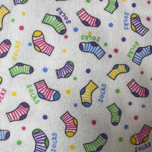 Może przedstawiać: Biały materiał z powtarzającym się wzorem kolorowych skarpet. Skarpety są w paski i mają różne kolory, w tym czerwony, niebieski, zielony, żółty i fioletowy. Słowo "skarpetki" jest nadrukowane na materiale czarnym kolorem.