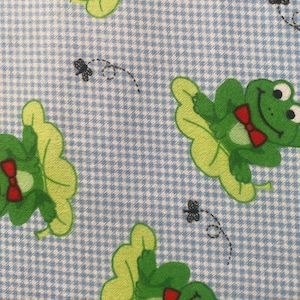 May include: A blue and white gingham fabric with a repeating pattern of green frogs wearing red bow ties sitting on lily pads.  The frogs have large eyes and are smiling.  There are also small black flies flying around the frogs.