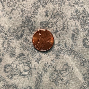 Może przedstawiać: Moneta amerykańska o nominale jednego centa, wykonana z miedzi, z widocznymi napisami "LIBERTY" i "IN GOD WE TRUST". Moneta leży na białym tle z kwiatowym wzorem.
