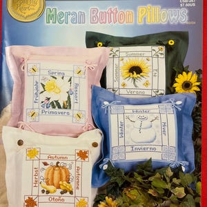 Puede incluir: Un folleto de patrones de punto de cruz para cuatro almohadas con botones, cada una representando una estación diferente. Las almohadas son de tela rosa, blanca, azul y verde. La portada del folleto presenta el título "Meran Button Pillows" y el texto "Cross My Heart, Inc."