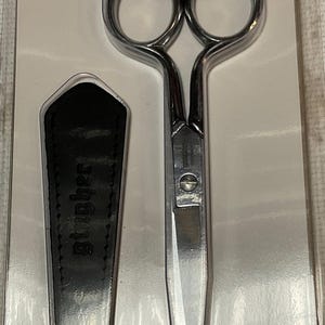 Puede incluir: Tijeras Gingher de color plateado con una funda de cuero negro. El embalaje incluye el nombre de la marca "Gingher" y el texto "una tradición de calidad". La etiqueta de precio dice "$29.99". El número de producto es "G-5".