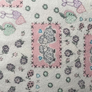 Może przedstawiać: Biała tkanina z powtarzającym się wzorem szarych owiec, różowych kwadratów, kwiatów i serc. Owieczki znajdują się w różowych kwadratach z czarnymi obramowaniami ściegiem krzyżykowym. Kwiaty i serca są w odcieniach fioletu, zieleni i błękitu.