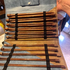 Peut inclure: Un ensemble d'aiguilles à tricoter en bois dans un étui en cuir bleu et beige. Les aiguilles sont de différentes tailles et maintenues par des sangles élastiques noires. L'étui est ouvert, présentant les aiguilles, et possède une fermeture à bouton.