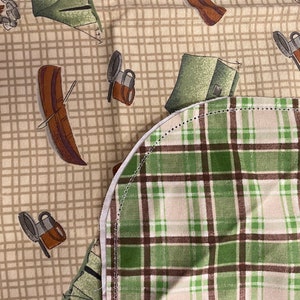 May include: A close-up of a fabric with a green and brown plaid pattern. The fabric is sewn with a white seam. The fabric is on top of a beige fabric with a pattern of camping equipment, including tents, canoes, and coffee mugs.