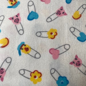 Może przedstawiać: Biały materiał z powtarzającym się wzorem szpilek bezpieczeństwa z różnokolorowymi zawieszkami. Zawieszki obejmują żółty kwiat, niebieskie serce, różowe serce i świnkę.