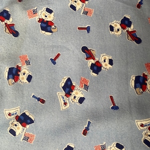 Może przedstawiać: Niebieski materiał z powtarzającym się wzorem białych kotów w niebieskich i czerwonych mundurach trzymających amerykańskie flagi. Koty trzymają czerwoną, białą i niebieską flagę z gwiazdą. Tło jest jasnoniebieskie.