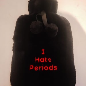 Pode incluir: Capa preta para garrafa de água quente com o texto bordado "I Hate Periods" em vermelho. A capa é feita de um material macio e felpudo e apresenta um topo fofo com dois pompons presos por uma fita preta.