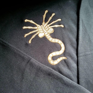 Può includere: Felpa nera con un design facehugger dorato ricamato. Il motivo raffigura la creatura aliena con il suo corpo allungato e molteplici appendici. Il design è centrato sul petto del capo di colore scuro.