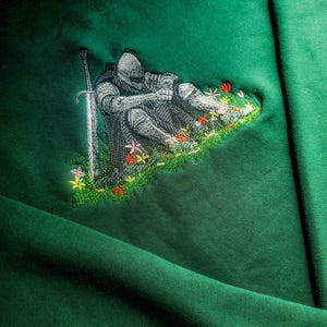May include: Emerald green sweatshirt featuring an embroidered design of a knight resting on a patch of grass with colorful flowers. A silver sword stands upright beside the knight. The design is detailed and the sweatshirt has a soft, textured appearance.