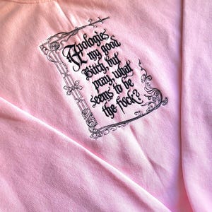 Puede incluir: Sudadera rosa claro con la frase "Apologies my good Bitch, but pray, what seems to be the f*ck?" en negro con una fuente decorativa. El texto está enmarcado por un borde negro ornamentado.