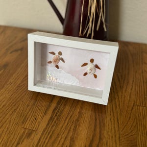 May include: A white framed shadow box featuring two sea turtle designs made from seashells. The turtles are set against a pink background with a white wave design. The frame is sitting on a wooden surface.