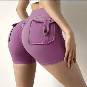 May include: Lavender-colored athletic shorts with high waist and pocket details. The shorts feature a fitted design with a button closure on the pockets. The fabric appears to be a stretchy, athletic material.