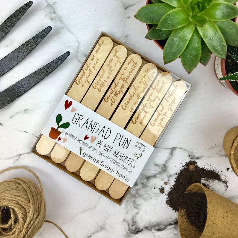 May include: A set of six wooden plant markers with humorous puns about grandads. The markers are packaged in a clear plastic box with a white label that reads "Grandad Pun Plant Markers" and "6 Amusing Expressions of Love for Green Fingered Grandads".