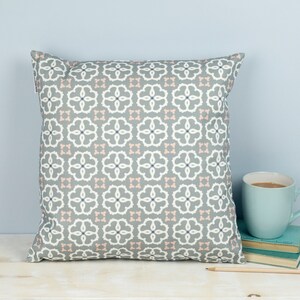 May include: A gray and white patterned throw pillow with pink accents. The pillow is on a light wood surface with a blue mug and books.