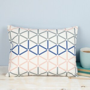 Puede incluir: Una almohada blanca con un patrón geométrico en tonos grises, azules y rosas. El patrón está formado por triángulos y cuadrados.