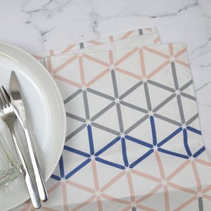 May include: A white cotton kitchen towel with a geometric pattern in shades of pink, gray, and blue. The pattern is made up of triangles and squares.