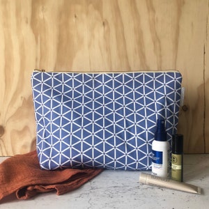 May include: A blue and white geometric patterned zippered pouch with a gold zipper. The pouch is sitting on a light brown wooden surface with a brown cloth and three small bottles.