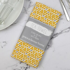 Peut inclure: Une serviette de th&eacute; jaune et grise avec un motif g&eacute;om&eacute;trique. La serviette est pli&eacute;e et porte une &eacute;tiquette sur laquelle est &eacute;crit "Tea Towel Designed and Made in the United Kingdom grace & favour home".