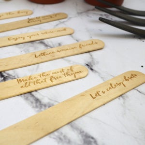 May include: Wooden plant markers with handwritten messages, including "Enjoy your nature-mint", "Lettuce romaine friends", "Make the most of all that free thyme", and "Let's celery-brate".