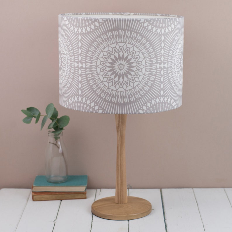 May include: A gray table lamp with a white patterned lampshade. The lampshade has a mandala design. The lamp is on a white table with a stack of books and a vase with greenery.