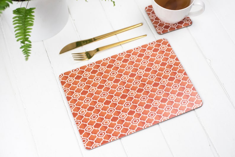 May include: Orange and white geometric patterned placemat with gold silverware on a white table.