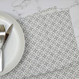 May include: A white ceramic plate with a fork and knife set on a gray patterned placemat. The placemat features a repeating geometric design in grey on a white background. The setting is on a marble surface.
