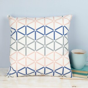Puede incluir: Una almohada blanca con un patrón geométrico en tonos rosa, azul y gris. El patrón está formado por triángulos y cuadrados.