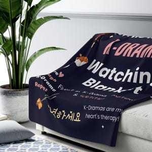 K-Drama Watching Blanket draped over a couch, showcasing fun K-Cozy K-Drama blanket with quotes like "Live Laugh K-Drama Repeat" and "K-Dramas are my heart's therapy!" Perfect for fans, surrounded by comfy decor.