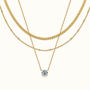 May include: A layered gold necklace set consisting of three distinct chains. The top chain is a thick curb link, the middle is a delicate beaded chain, and the bottom chain features a single round brilliant-cut clear stone pendant.