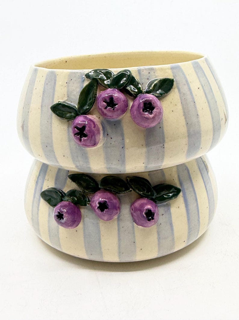 May include: Two stacked ceramic bowls with a cream and light blue striped pattern. Each bowl is decorated with three purple blueberry clusters with green leaves. The bowls have a rounded shape and a light beige interior.