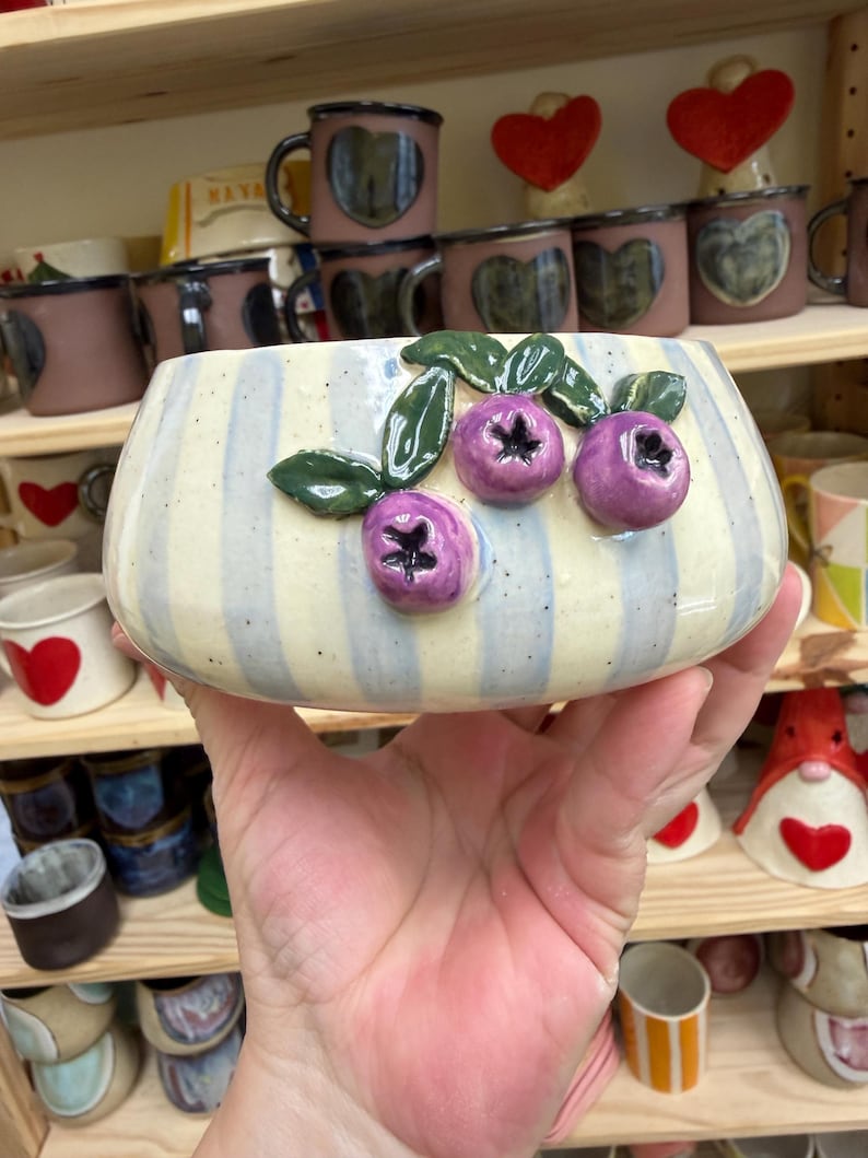 May include: A ceramic bowl with a cream and blue striped pattern, adorned with three purple blueberry sculptures and green leaves. The bowl is held in a hand, with other ceramic items visible on shelves in the background.