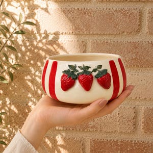 May include: A ceramic bowl with a cream-colored base and red vertical stripes. Three raised, red strawberries with green leaves are centered on the bowl. The bowl is held in a person's hand.