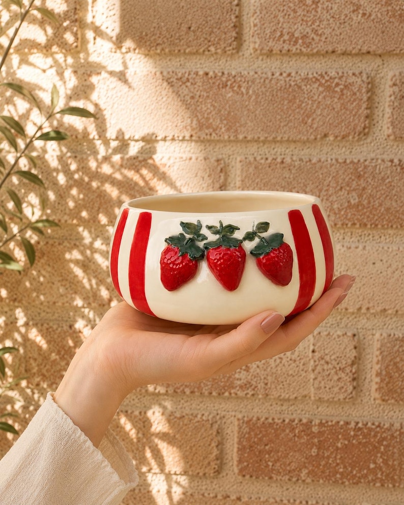May include: A ceramic bowl with a cream-colored base and red vertical stripes. Three raised, red strawberries with green leaves are centered on the bowl. The bowl is held in a person's hand.