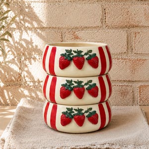 May include: A stack of three ceramic bowls. Each bowl is cream-colored with vertical red stripes and three raised red strawberry designs with green leaves. The bowls are stacked on a beige linen cloth.