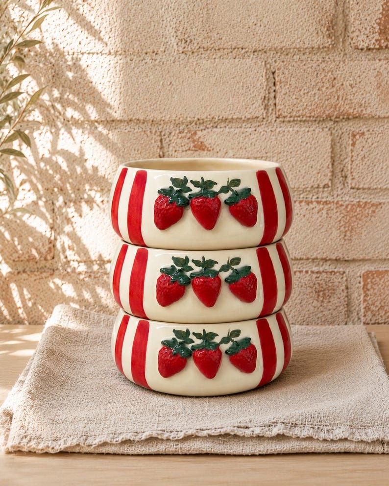 May include: A stack of three ceramic bowls. Each bowl is cream-colored with vertical red stripes and three raised red strawberry designs with green leaves. The bowls are stacked on a beige linen cloth.