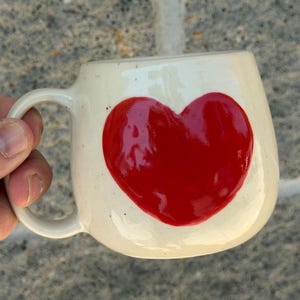 May include: Cream-colored ceramic mug with a large, glossy red heart design. The mug has a rounded shape and a curved handle. The background is a textured gray surface.
