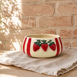 May include: A cream-colored ceramic bowl with red vertical stripes and three raised strawberry designs. The strawberries are red with green leaves. The bowl sits on a beige linen cloth.