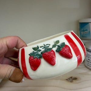 May include: A ceramic bowl with a cream-colored base and red vertical stripes. The bowl features three raised, bright red strawberries with green leaves. The bowl is held in a person's hand.