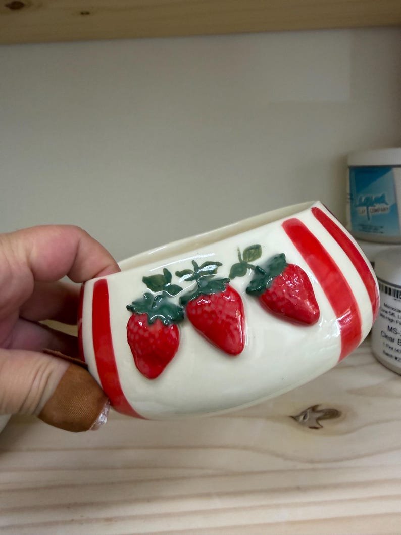 May include: A ceramic bowl with a cream-colored base and red vertical stripes. The bowl features three raised, bright red strawberries with green leaves. The bowl is held in a person's hand.