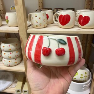 May include: A ceramic bowl with a white base and red stripes, featuring two red cherry accents. Other ceramic mugs and bowls with cherry and heart designs are visible on wooden shelves.