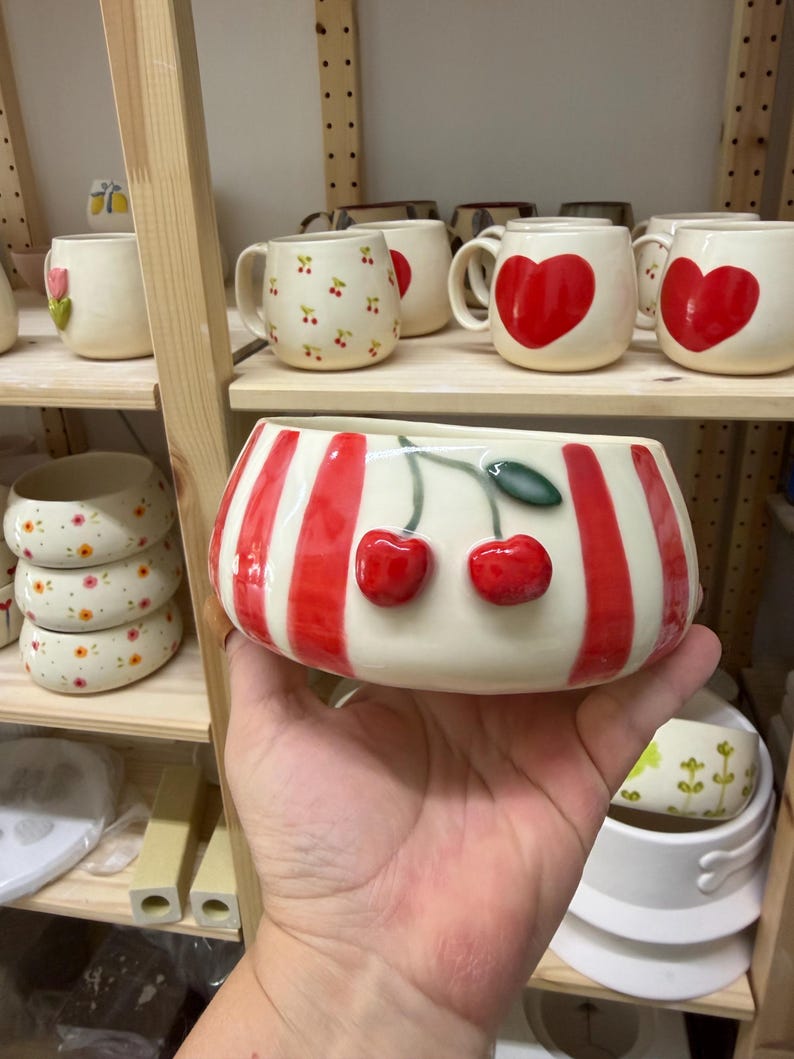 May include: A ceramic bowl with a white base and red stripes, featuring two red cherry accents. Other ceramic mugs and bowls with cherry and heart designs are visible on wooden shelves.