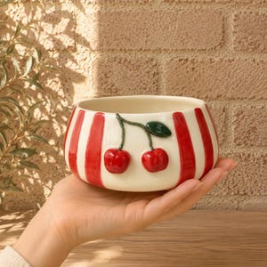 May include: A ceramic bowl with a cream base and vertical red stripes. Two red cherries with green leaves are on the side. The bowl is held in a person's hand. The background is a brick wall.