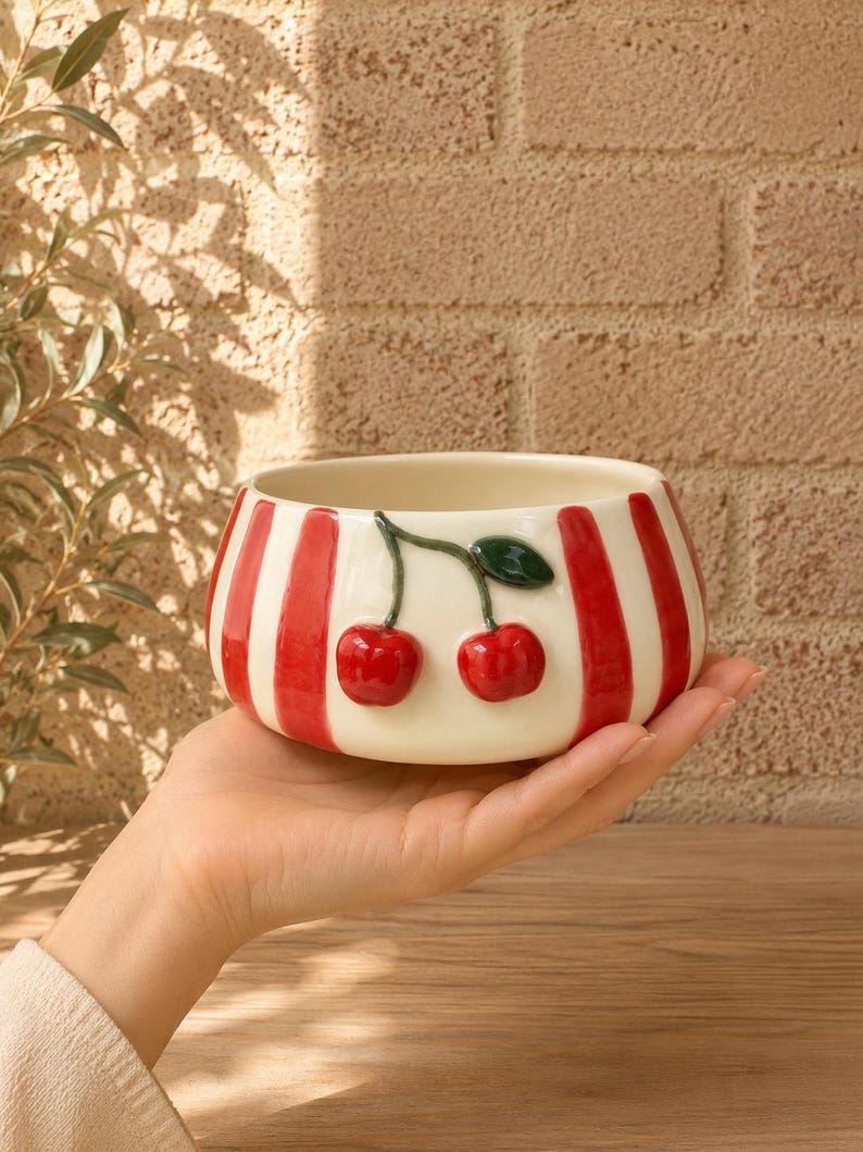 May include: A ceramic bowl with a cream base and vertical red stripes. Two red cherries with green leaves are on the side. The bowl is held in a person's hand. The background is a brick wall.